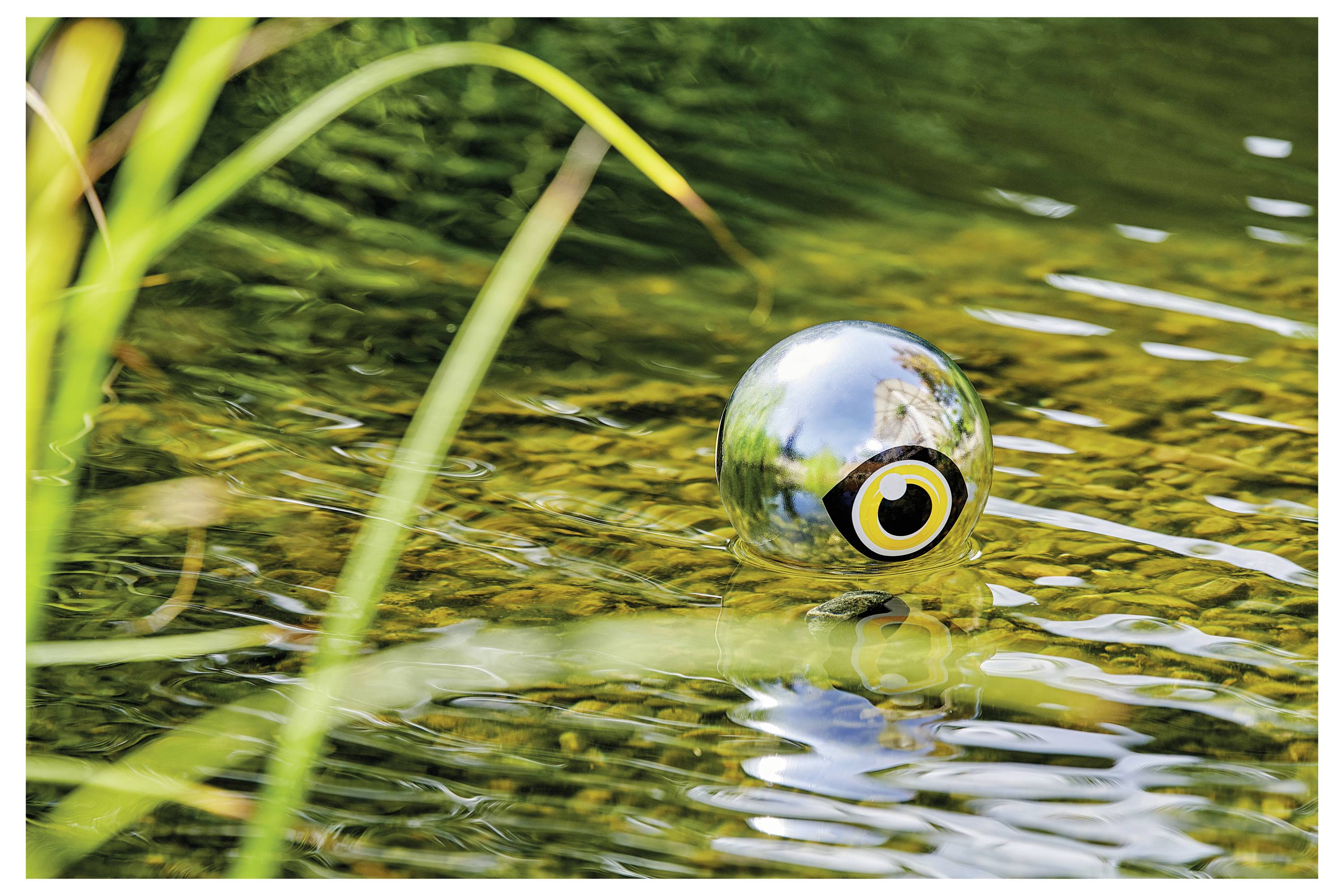 Refleksyjna srebrzysta kula z wzorem oka unosi się na stawie, otoczona wodą i zielenią, tworząc surrealistyczny efekt wizualny.