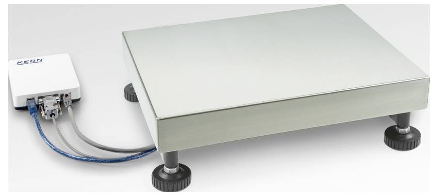 Prostokątna elektroniczna waga metalowa z czterema regulowanymi na wysokość stopkami oraz przewodem połączeniowym prowadzącym do białego urządzenia sterującego po lewej stronie.