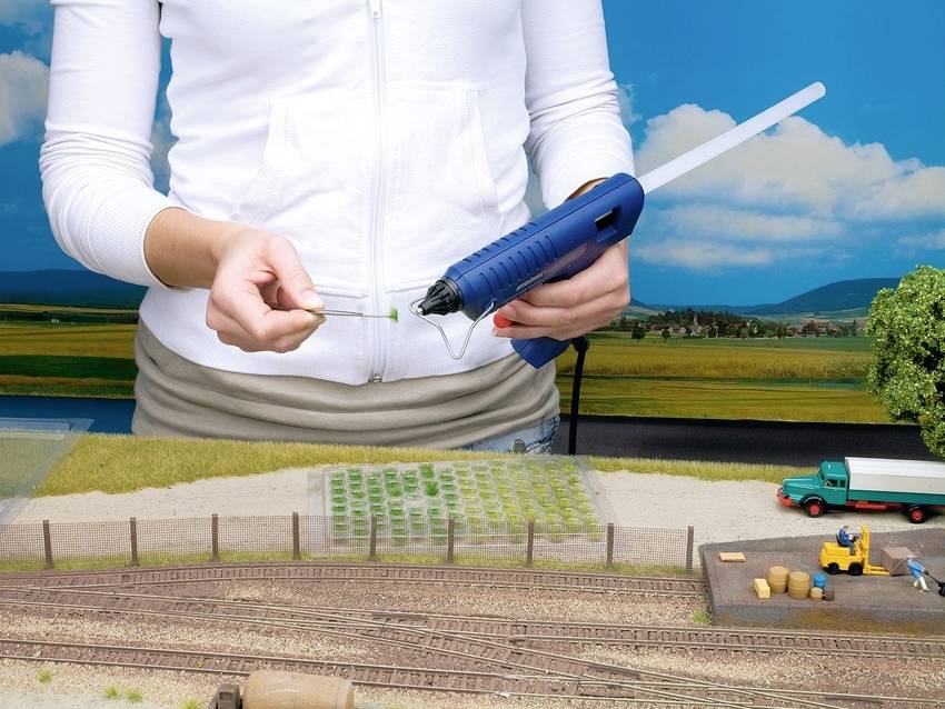 Osoba przyklejająca coś pistoletem do kleju na gorąco w miniaturowej scenerii. W tle widoczne są pola i tory modelarskiej kolejki.