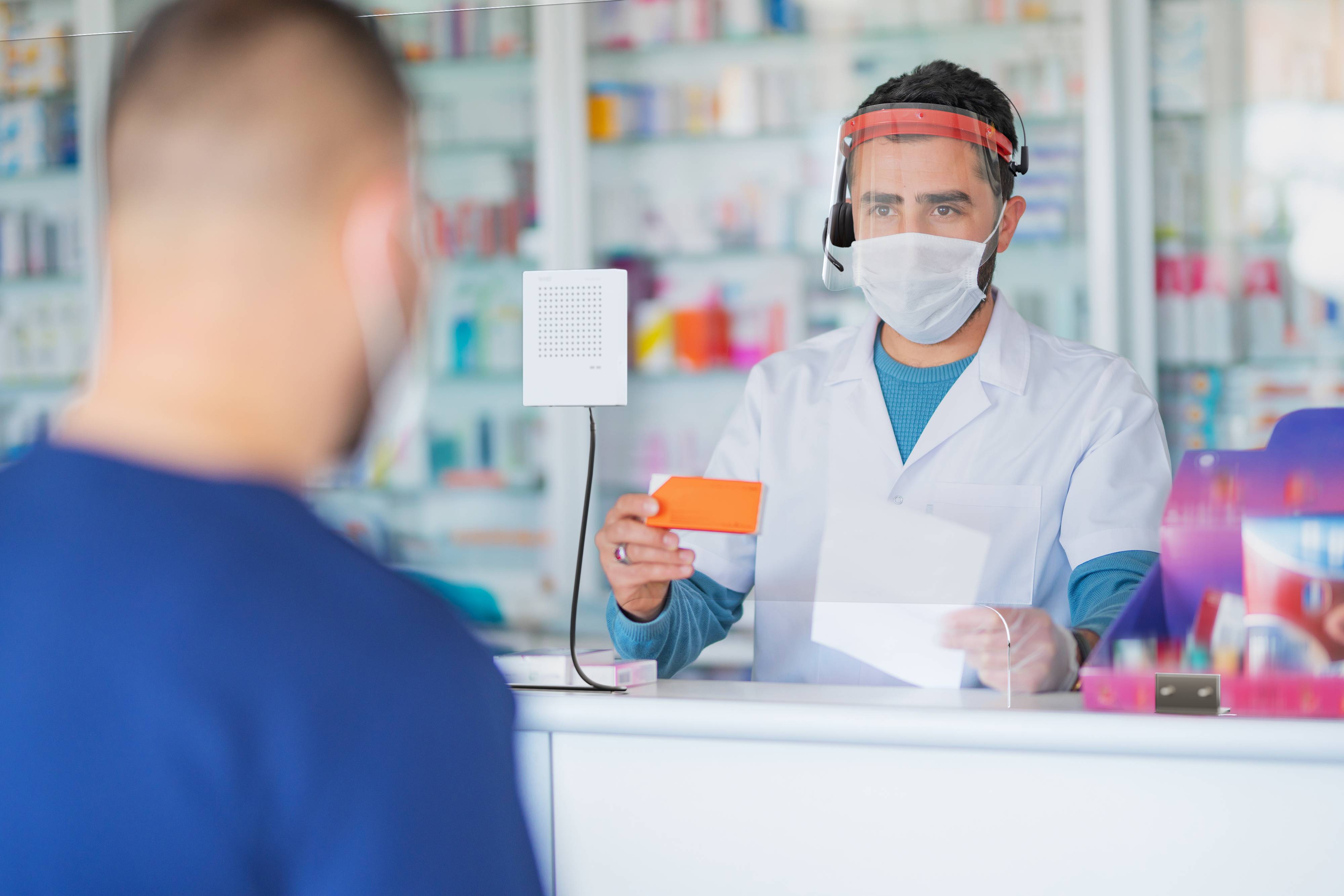 Farmaceuta za szybą z pleksiglasu, w masce i przyłbicy, wydaje klientowi leki i receptę.