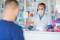 Farmaceuta za szybą z pleksiglasu, w masce i przyłbicy, wydaje klientowi leki i receptę.