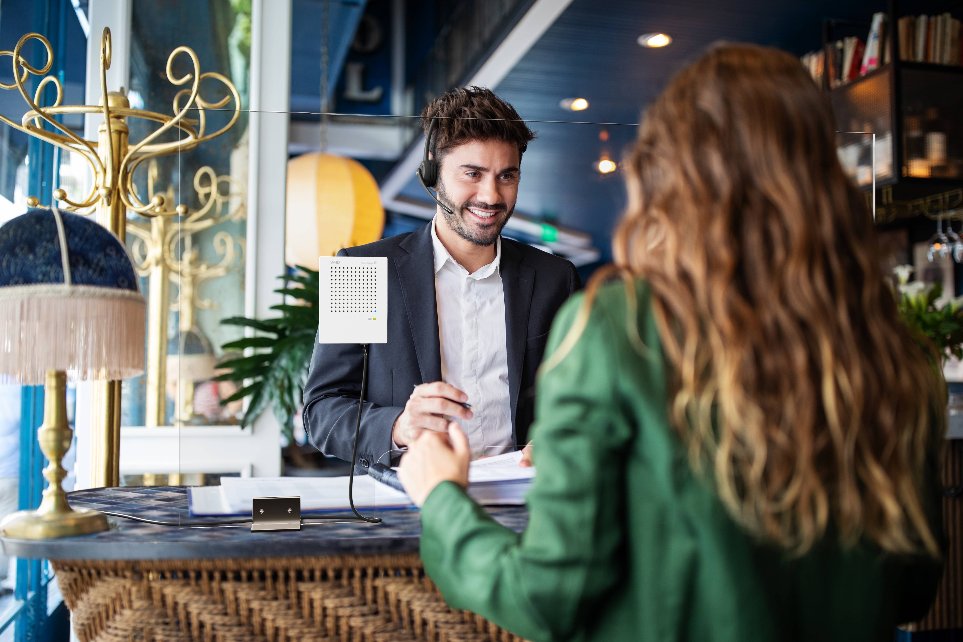 Mężczyzna za ladą recepcyjną uśmiecha się, rozmawiając z kobietą. Ma na głowie zestaw słuchawkowy i trzyma w ręku dokumenty.