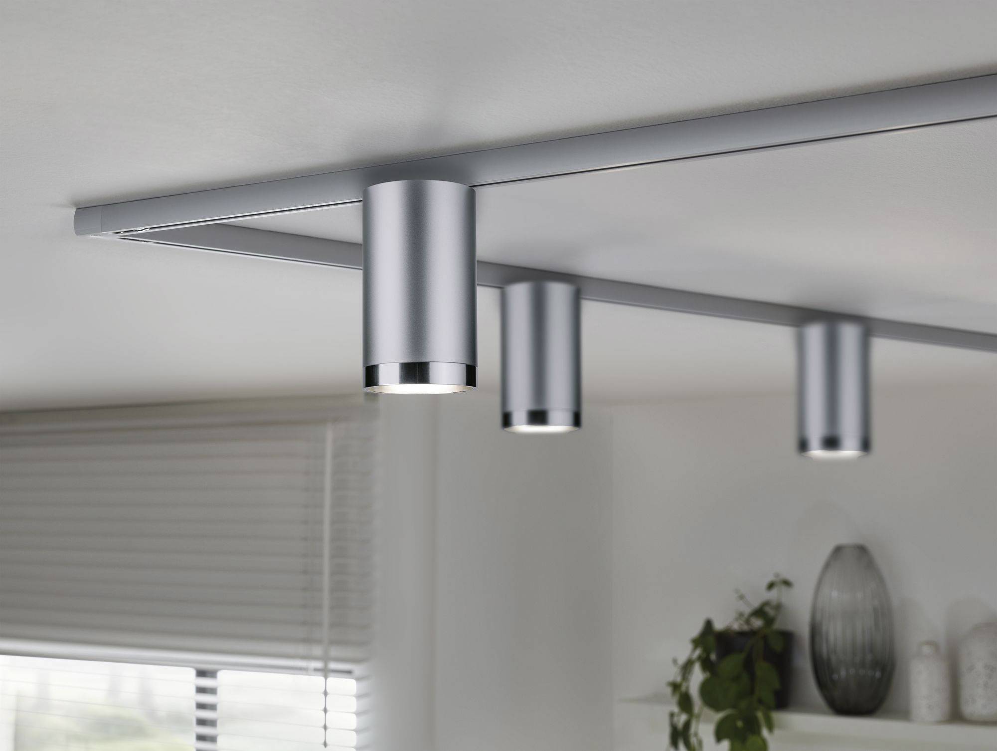 Srebrne lampy sufitowe świecą w nowoczesnym pokoju mieszkalnym. Po lewej stronie znajduje się okno z żaluzjami, po prawej rośliny i dekoracje.