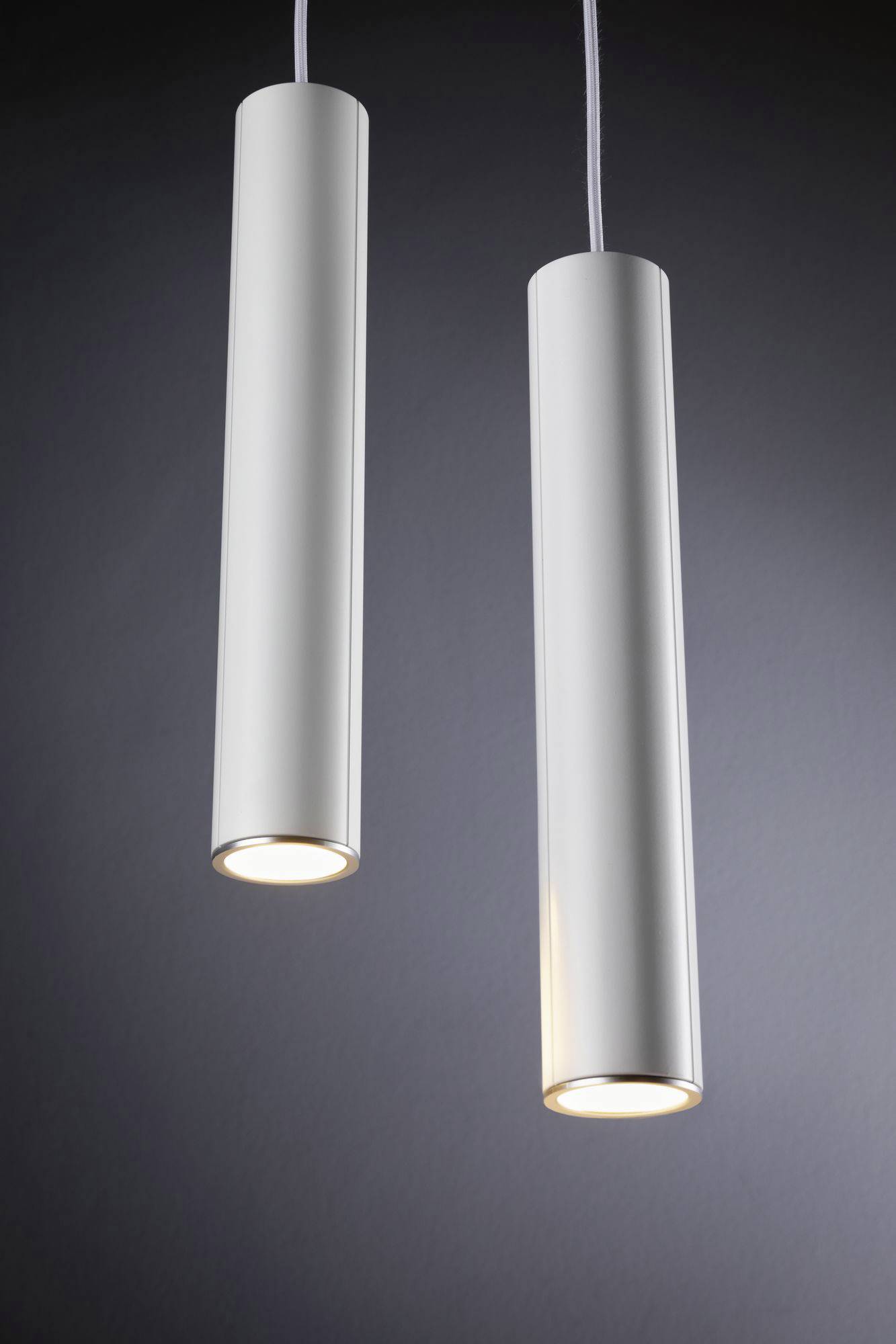 Dwie cylindryczne lampy wiszące zwisają z sufitu na ciemnym tle, świecą w dół i wytwarzają ciepłe światło.