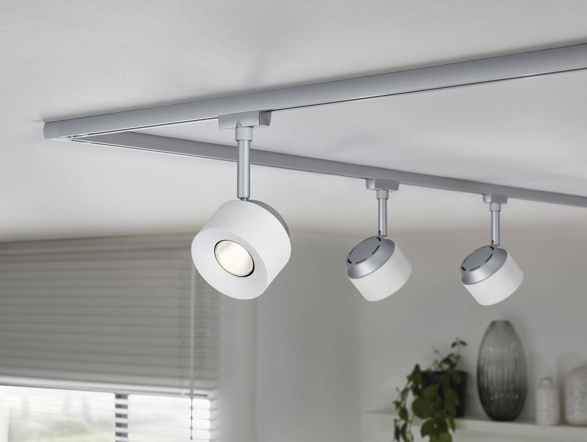 Trzy nowoczesne reflektory sufitowe, które można indywidualnie regulować, zamocowane są na srebrnej szynie i oświetlają pomieszczenie z roślinami i oknem.