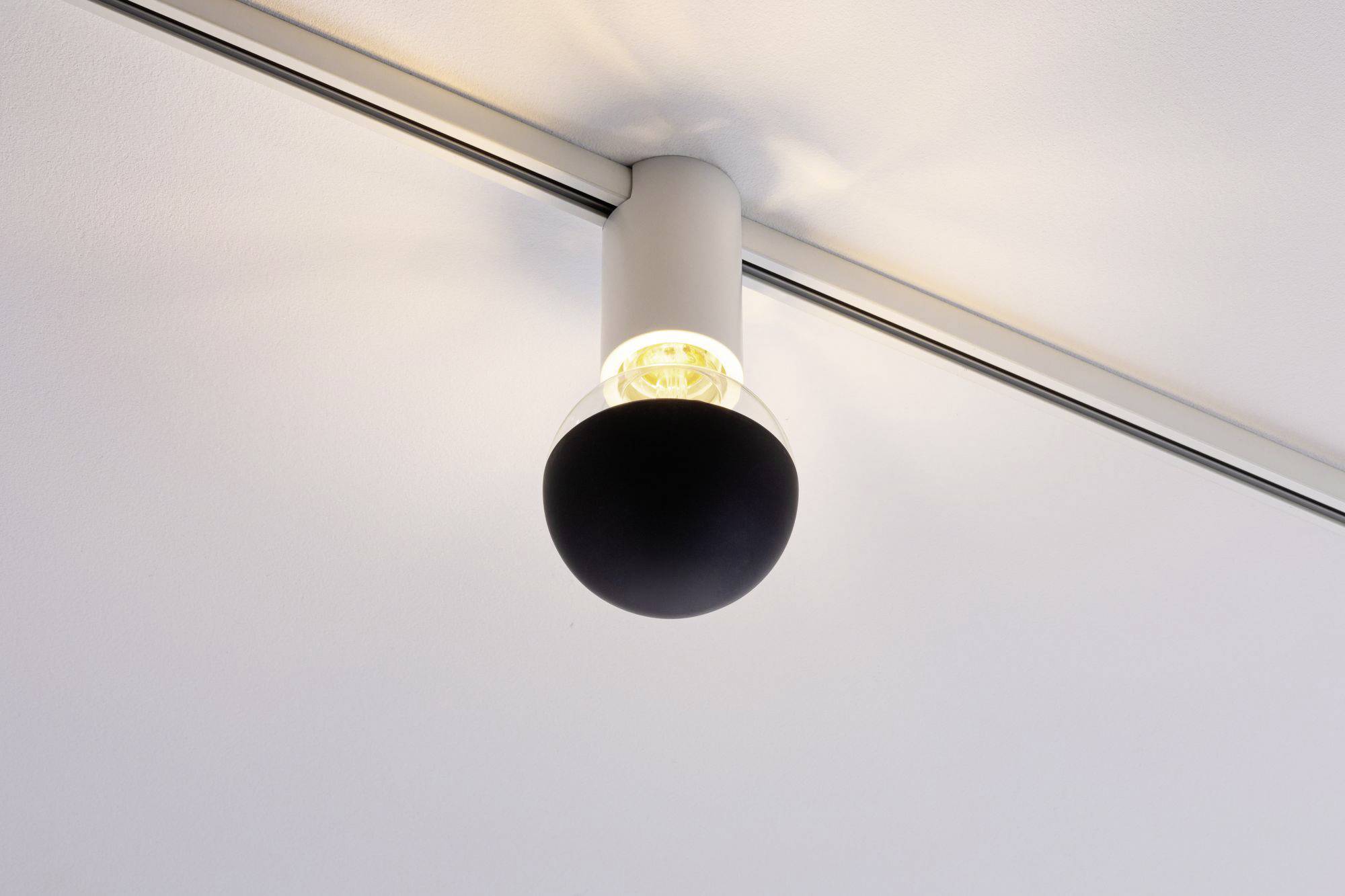 Nowoczesna lampa sufitowa z czarnym kloszem emituje ciepłe światło. Jest zamontowana na metalowej szynie.