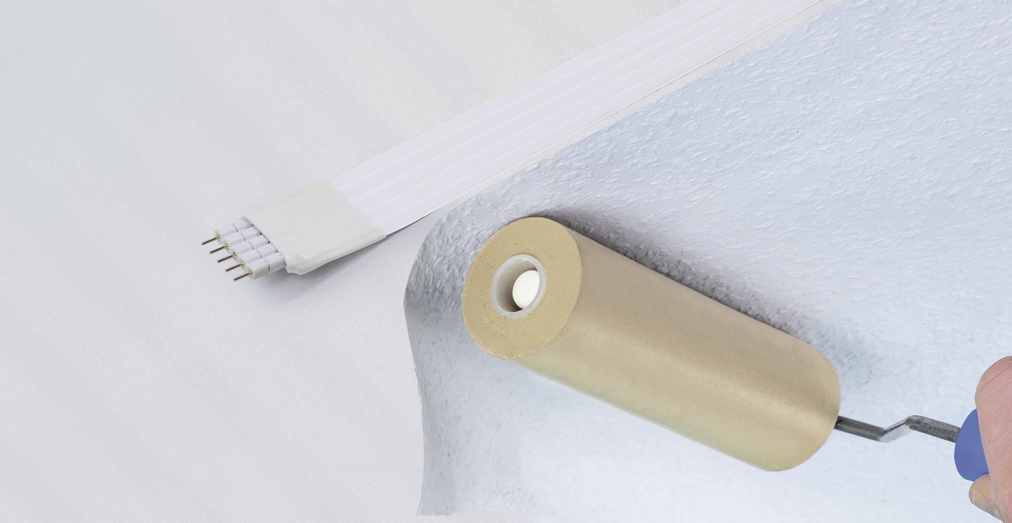 Dłoń trzyma wałek i przykłada jasnoniebieski fragment tapety z białym elementem łączącym do ściany.