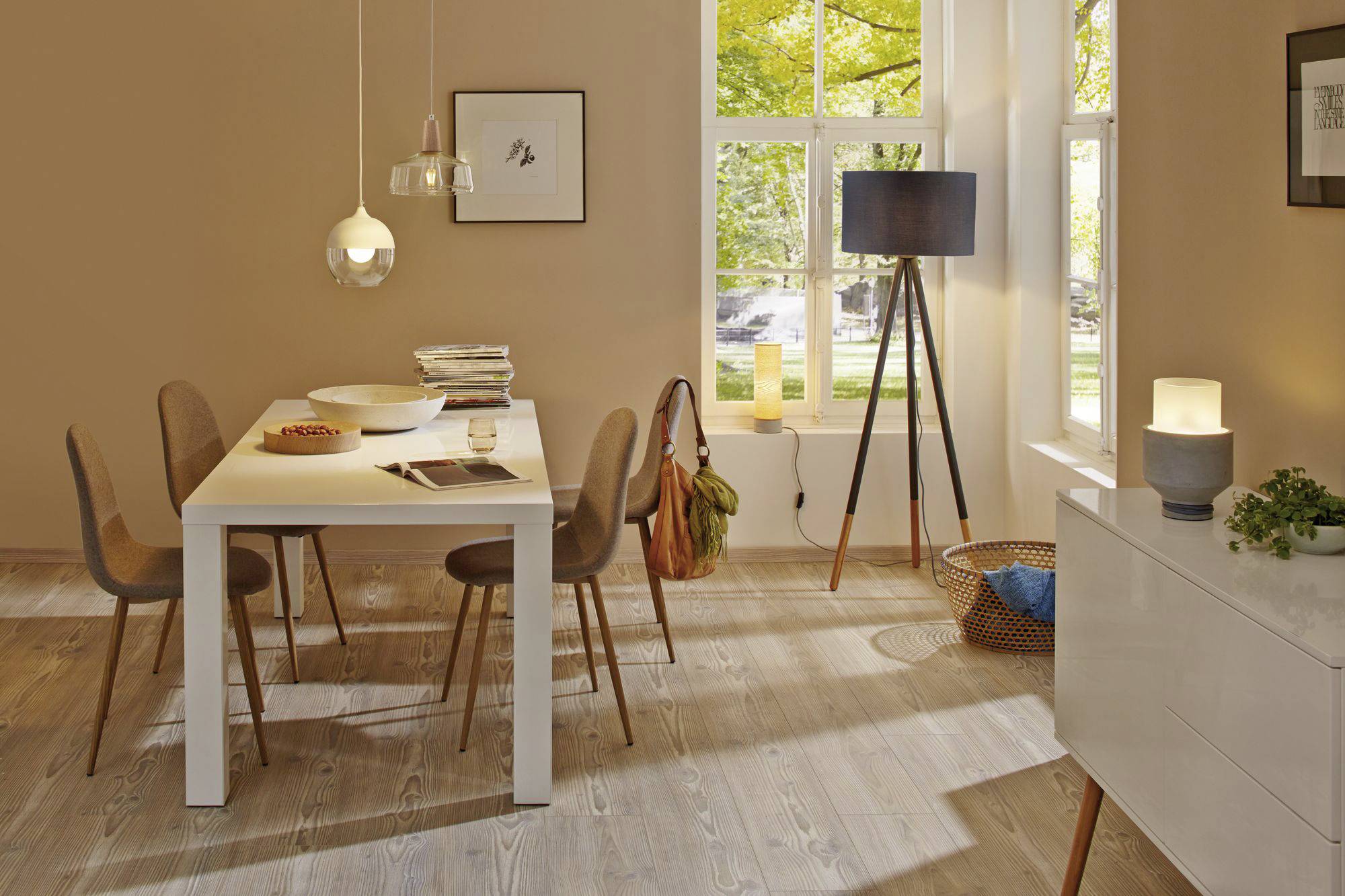 Jadalnia z drewnianą podłogą, białym stołem, sześcioma brązowymi krzesłami, lampami, oknem z widokiem na zieleń i komodą.