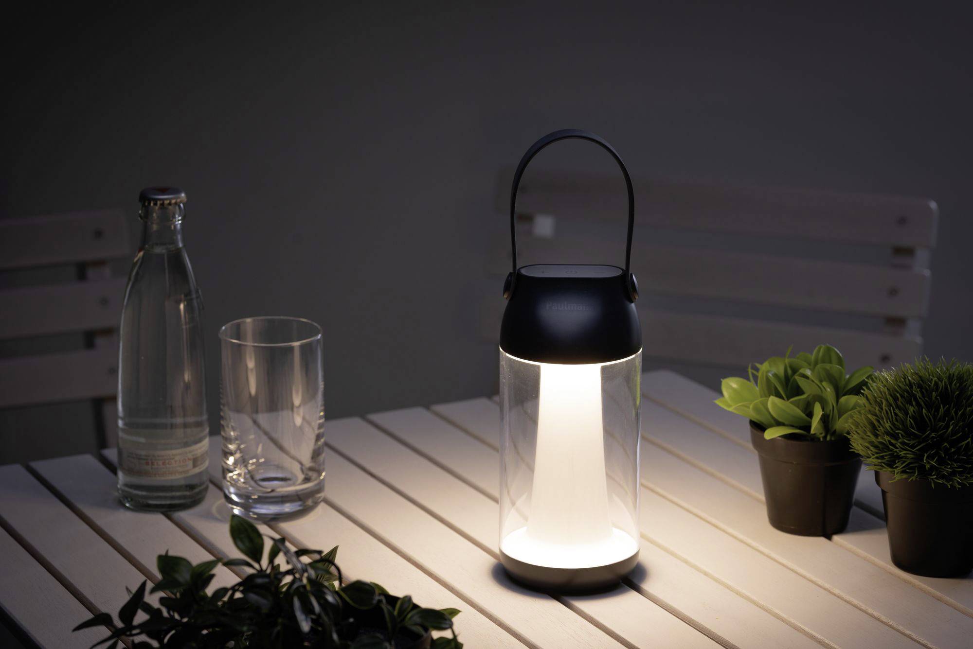 Na stole stoi świecąca latarnia obok butelki, szkła i małych roślin. Scena jest słabo oświetlona.