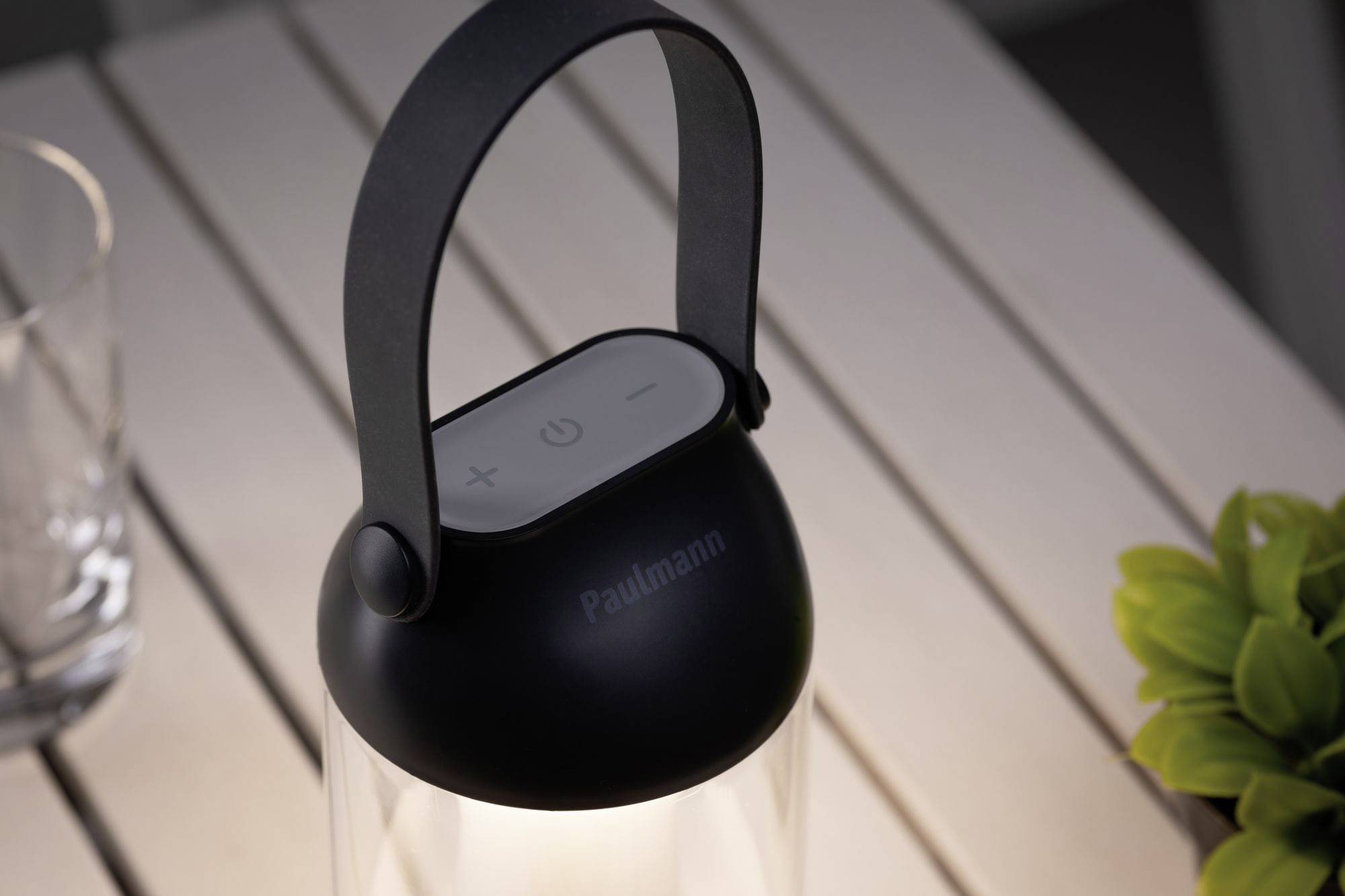 Przenośna lampa z uchwytem na drewnianym stole. Lampa posiada przyciski do regulacji jasności. Po prawej stronie zielony liść.