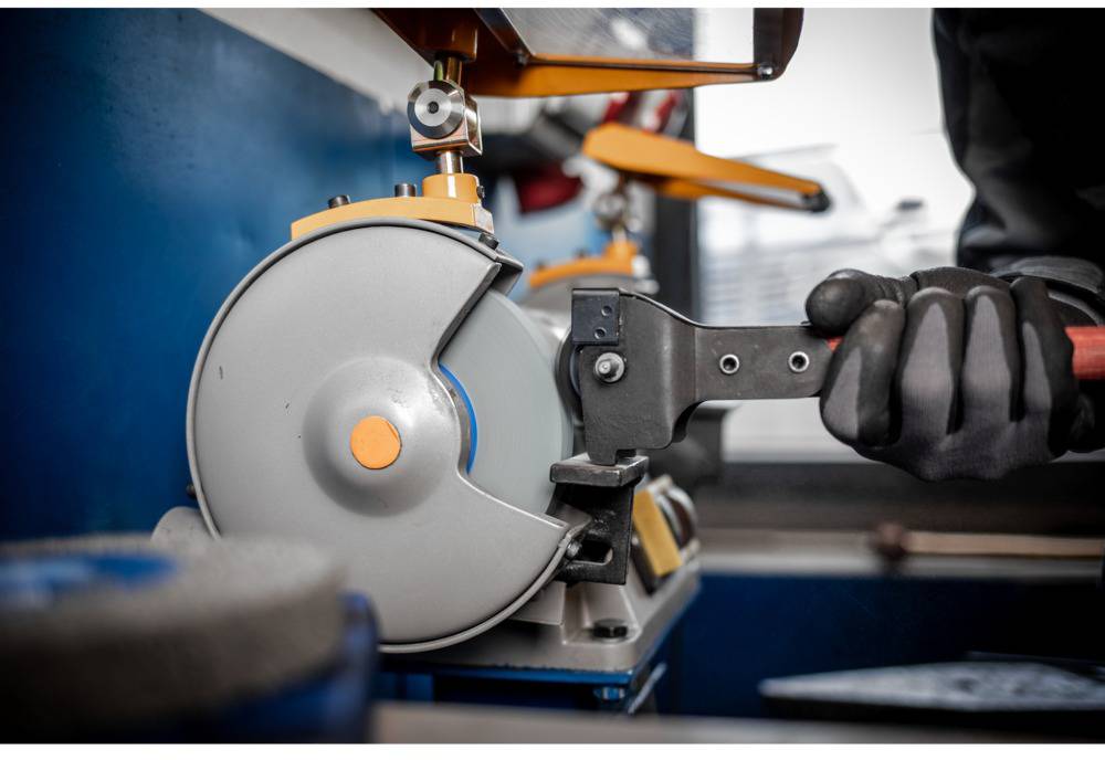 Robotnik szlifuje narzędzie przy użyciu szlifierki. Ma na rękach ciemne rękawice robocze. W tle widoczne są inne narzędzia.