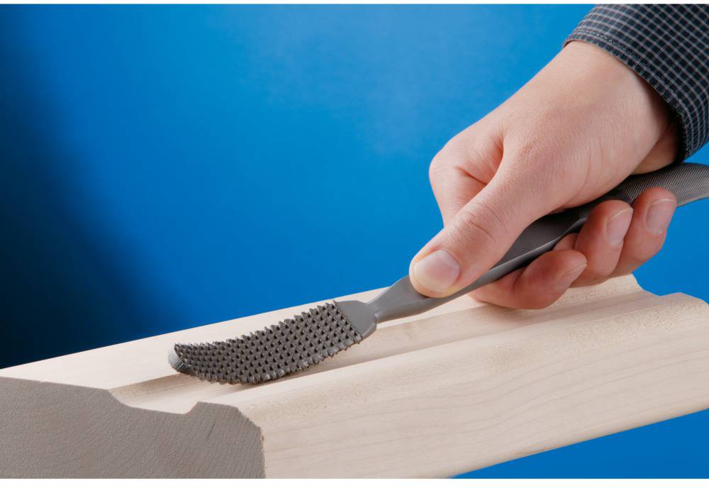 Dłoń trzyma tarnik i obrabia kawałek drewna na niebieskim tle.