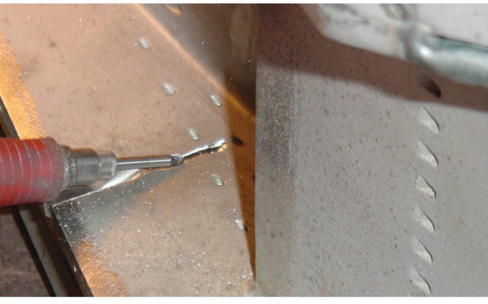 Zbliżenie obracającej się wiertarki, która wykonuje otwór w blasze metalowej. Obok wiertła widoczne są wióry metalowe.