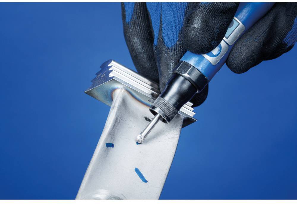 Dłoń w rękawicy trzyma srebrny element metalowy i używa niebieskiego narzędzia do usuwania zadziorów z krawędzi. Tło jest niebieskie.