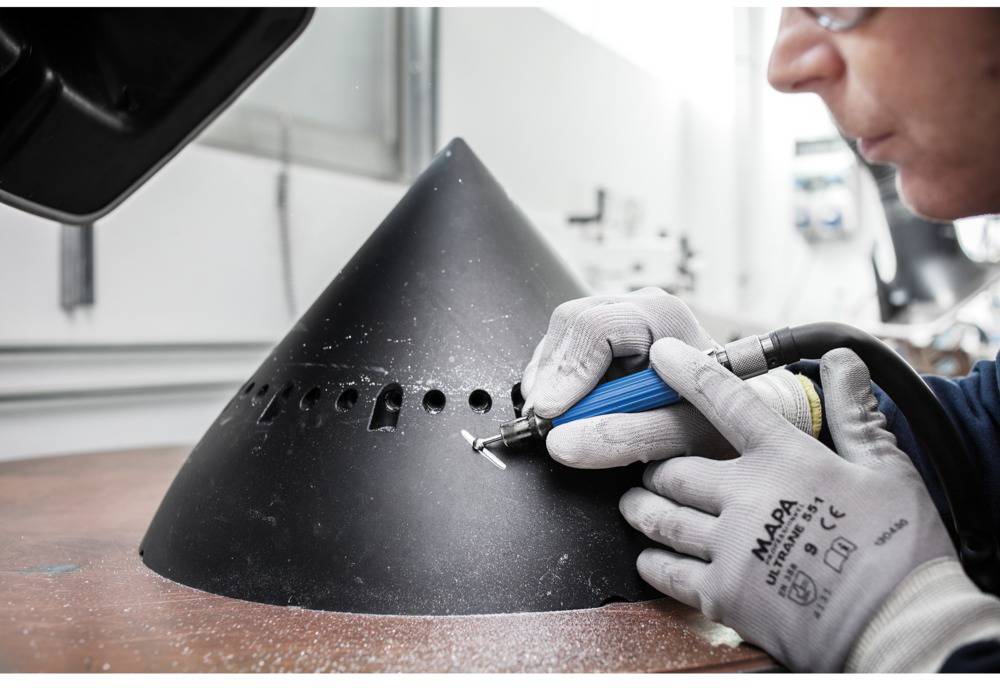 Jedna osoba obrabiarką elektryczną przetwarza dużą, czarną, stożkowatą formę metalową. Pracownik jest ubrany w rękawice ochronne i okulary zabezpieczające.