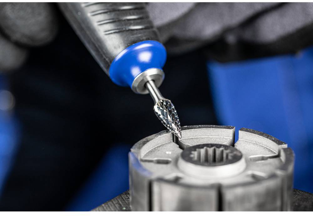 Zbliżenie narzędzia obrotowego z głowicą szlifierską obrabiającego metaliczny sworzeń zębaty. Na rozmytym tle widoczne są rękawice robocze.