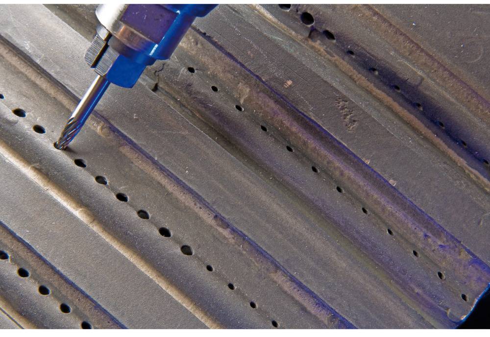 Zbliżenie wiertła drążącego otwory w falistym arkuszu blachy. Wiertarka jest niebieska.