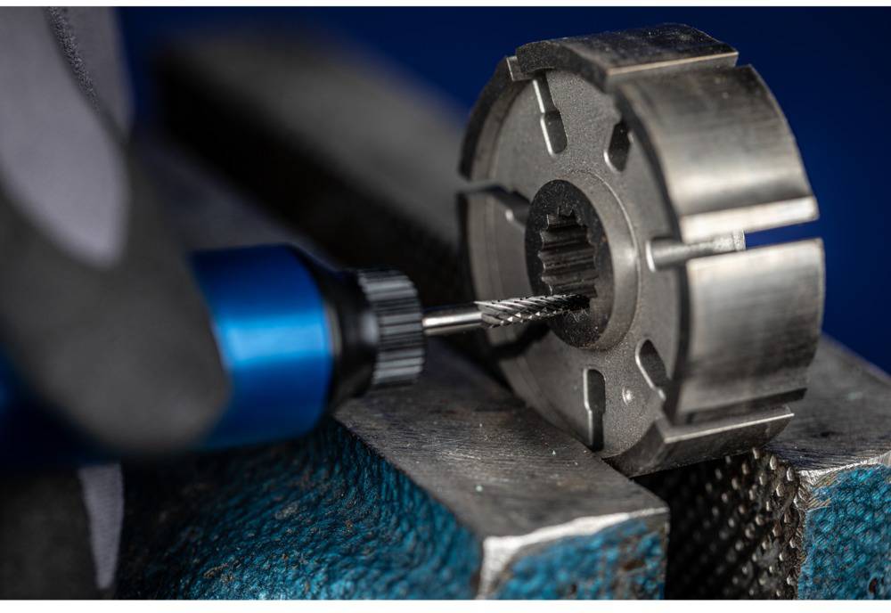 Wiertło obrabia metalowy element zamocowany w imadle. Ręka trzyma narzędzie, wykonując precyzyjną pracę.