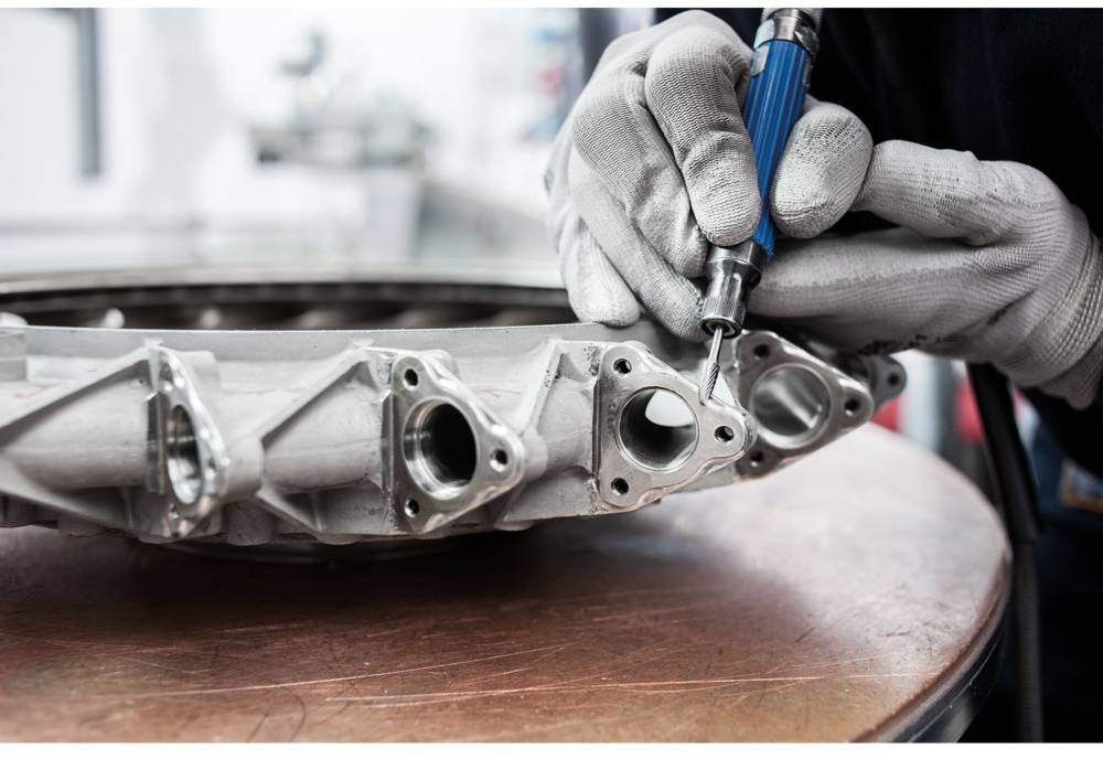 Osoba w rękawicach używa narzędzia do obróbki metalowego elementu maszyny na stole.