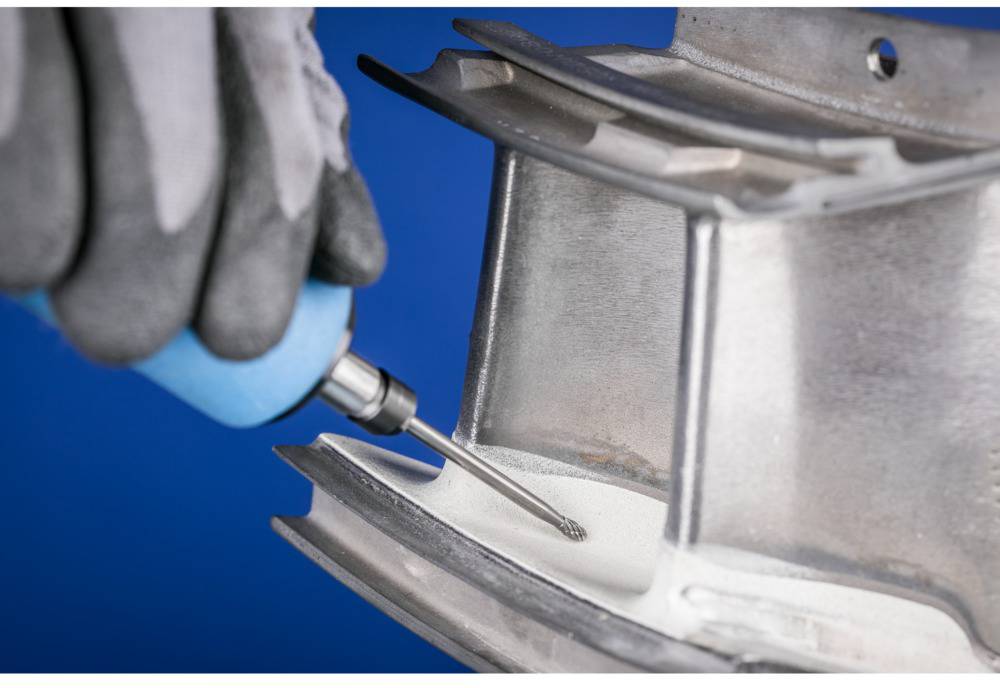 Osoba w rękawicach obrabia powierzchnię metalową obrotowym narzędziem, prawdopodobnie w celu polerowania lub szlifowania.