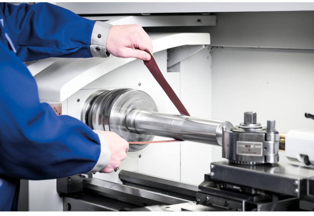 Pracownik pracuje przy tokarce i szlifuje cylindry metalowe za pomocą taśmy ściernej, ubrany w niebieskie ubranie robocze.