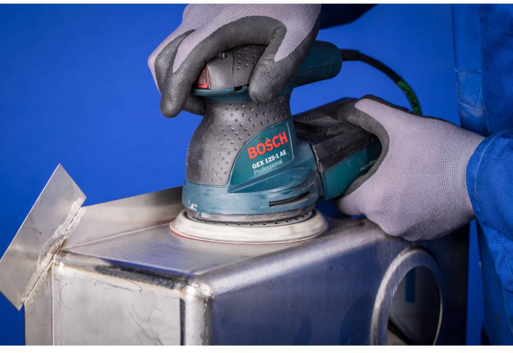 Osoba szlifuje metalową powierzchnię szlifierką mimośrodową Bosch GEX 125-1 AE. Osoba ma na rękach szare rękawice.