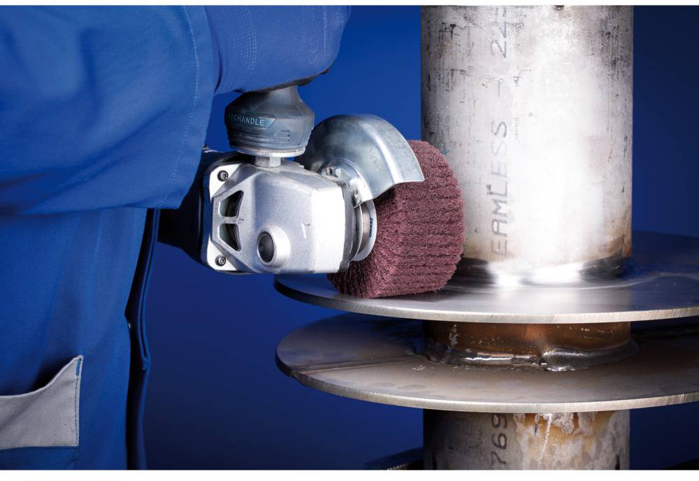 Osoba szlifuje rurę metalową za pomocą szlifierki kątowej. Ma na sobie niebieskie ubranie robocze. Rura jest zamocowana przy spawie.