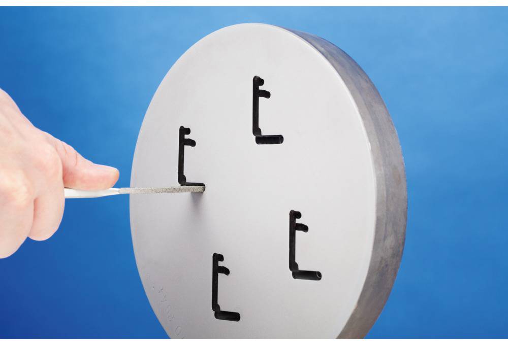Jedna ręka trzyma metalowe narzędzie, które jest wkładane w jeden z pięciu L-kształtnych wycięć na okrągłej metalowej płycie.