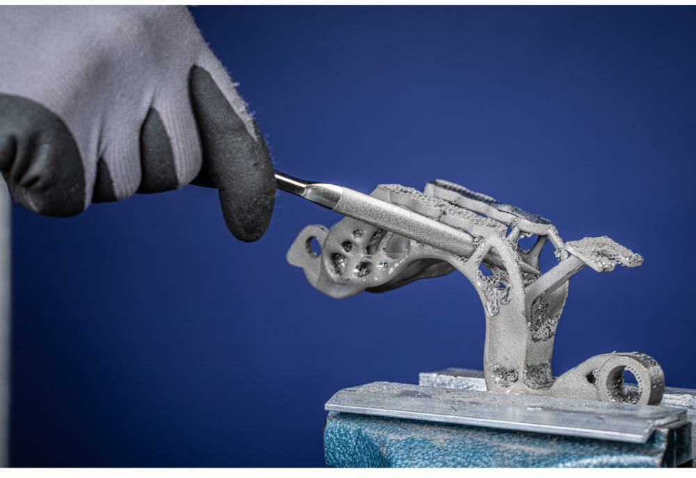 Osoba w rękawiczkach pracuje narzędziem przy skomplikowanej metalowej części wykonanej za pomocą drukarki 3D.