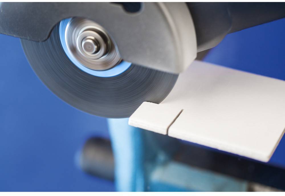 Piła do glazury ucina białą płytkę ceramiczną. Obracająca się tarcza tnąca dotyka płytki, a widoczne są niebieskie refleksy.