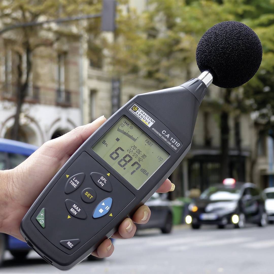 Jedna ręka trzyma miernik poziomu dźwięku wskazujący 68,7 dB, na miejskiej ulicy z przejeżdżającymi samochodami i drzewami w tle.