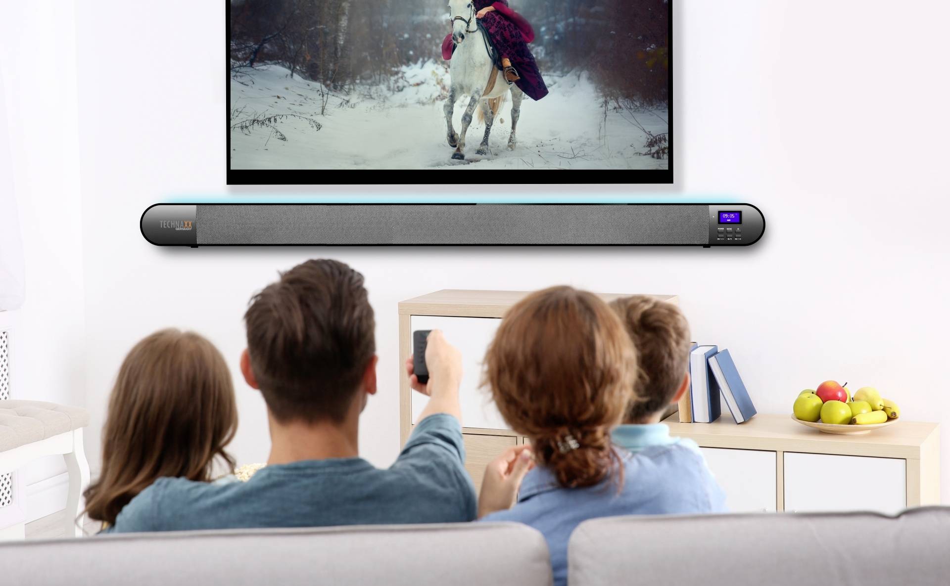 Rodzina ogląda film z sceną jeździecką na telewizorze; pod spodem znajduje się soundbar.