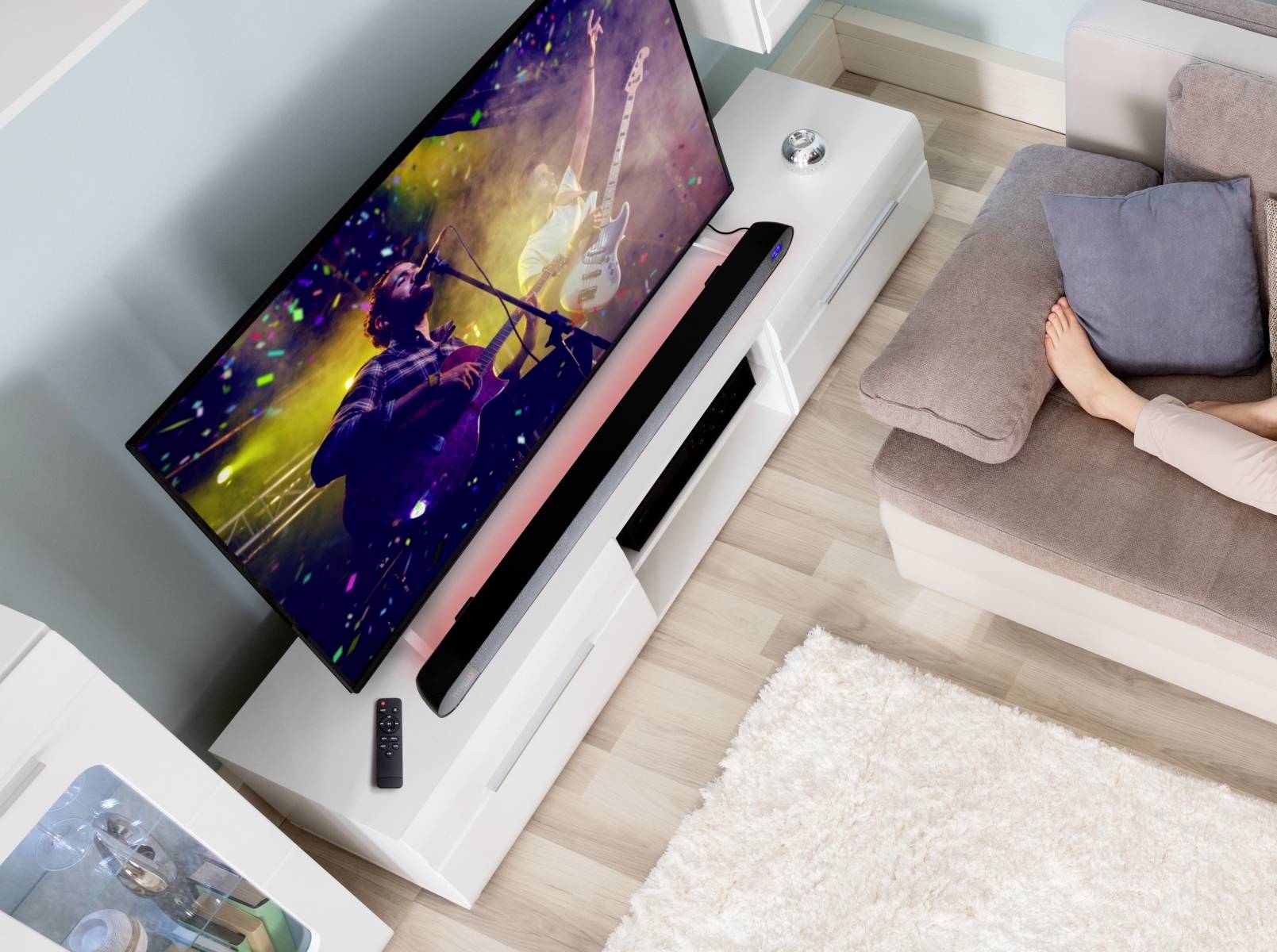 Telewizor wyświetla występ muzyczny, podczas którego jedna osoba gra na gitarze. Obok, na kanapie, siedzi inna osoba.