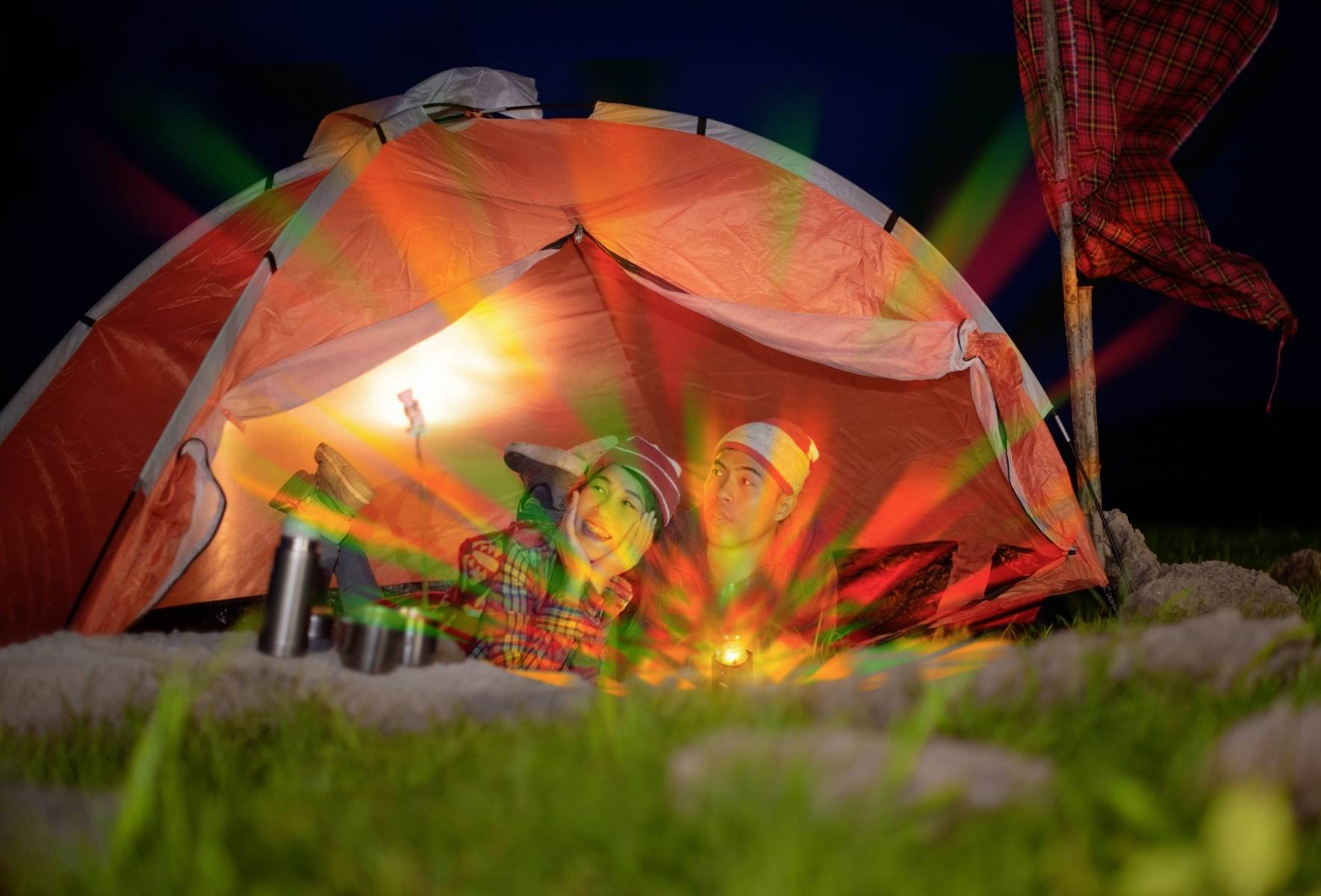 Dwie osoby leżą nocą w oświetlonym namiocie na łące, otoczone sprzętem kempingowym pod gwiaździstym niebem.
