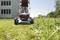 Osoba kosi trawę kosiarką Metabo przed budynkiem, otoczona drzewami i łąką w słoneczny dzień.