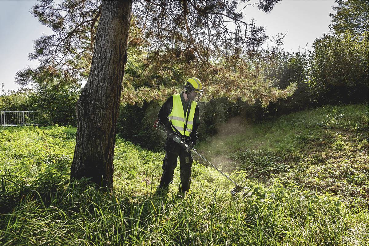 Osoba kosi wysoką trawę pod drzewem przy pomocy podkaszarki. Ma na sobie odzież ochronną, kamizelkę ostrzegawczą i kask.