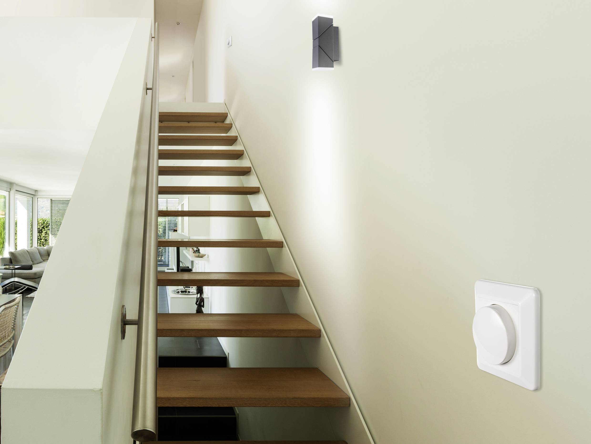 'Klatka schodowa z drewnianymi stopniami, kinkiet ścienny i ściemniacz na ścianie. W tle jasny salon.'