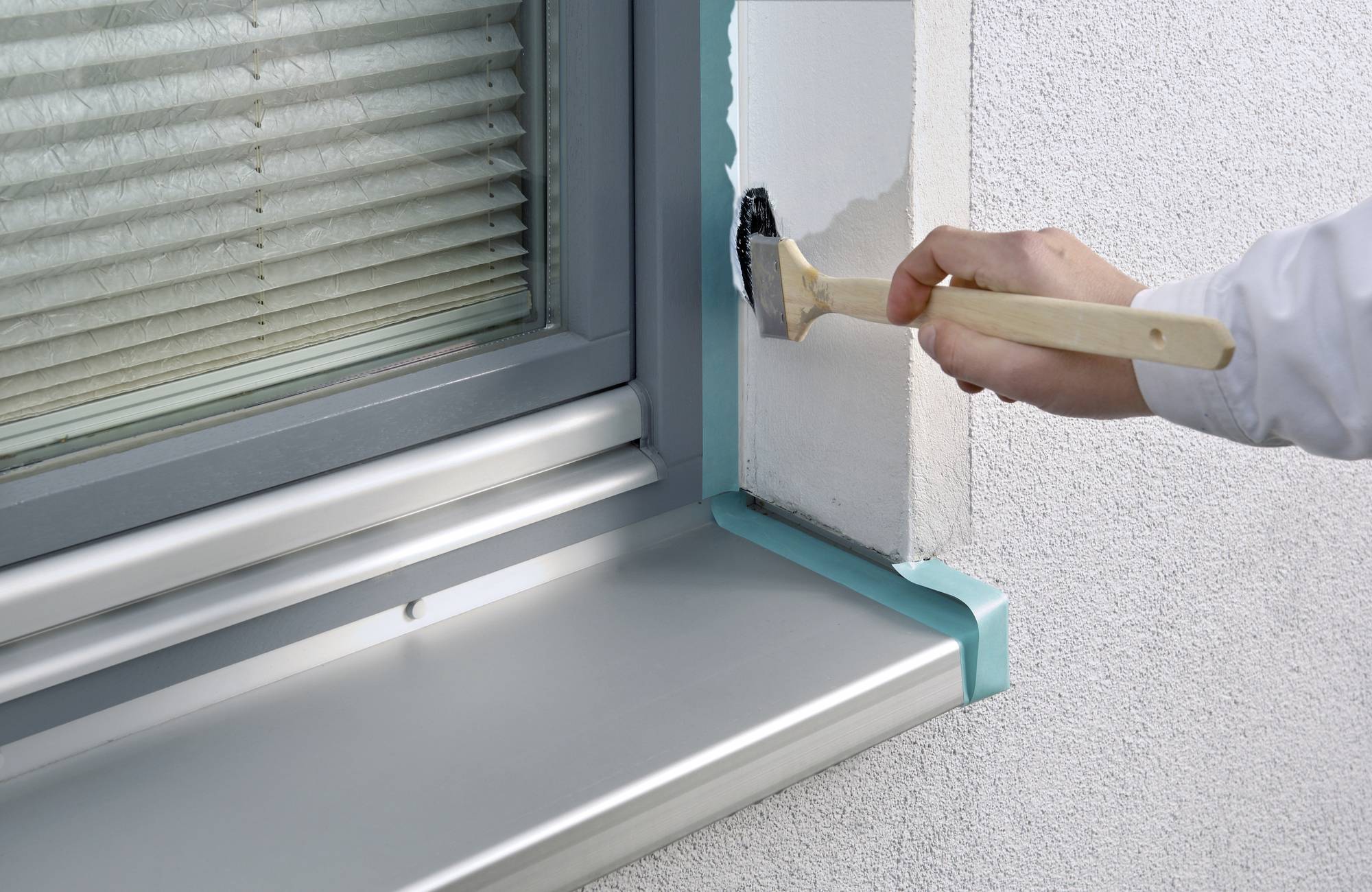 Ręka nakłada białą farbę na ścianę wokół ramy okiennej. Okno jest wyposażone w żaluzje, a rama jest częściowo zabezpieczona taśmą malarską.
