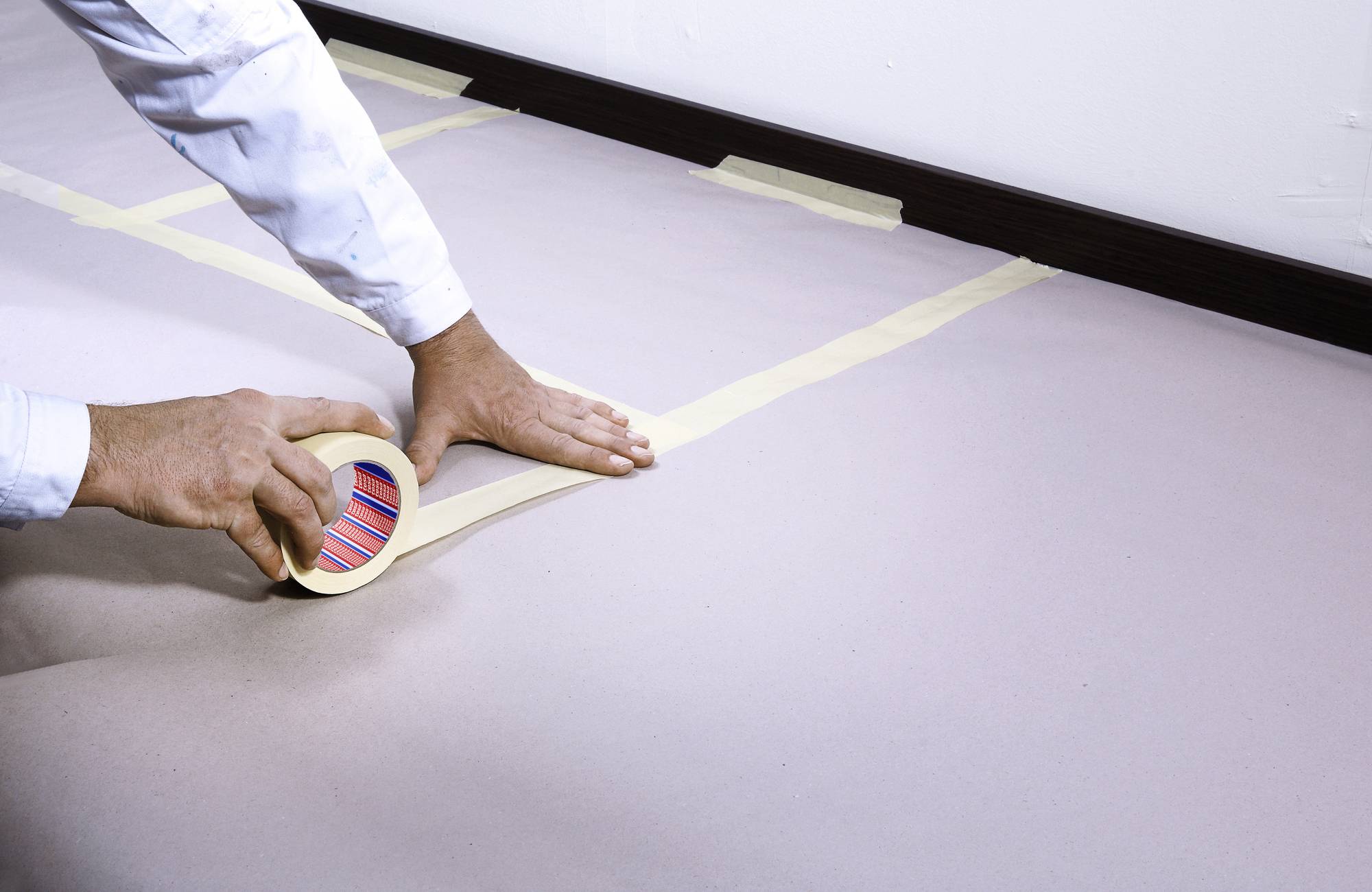 Osoba nakłada taśmę malarską na podłogę w pokoju, najprawdopodobniej w ramach przygotowań do prac malarskich lub zabezpieczenia podłogi.