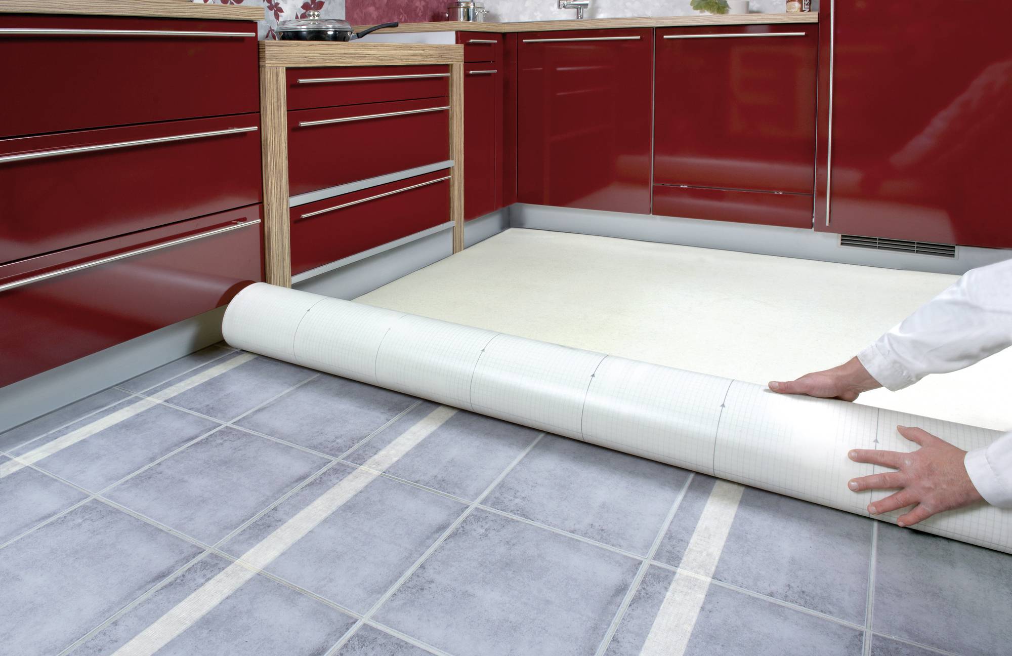 Osoba rozściela nowy jasny pokrycie podłogowe z winylu na wyłożonej płytkami podłodze kuchennej z czerwonymi szafkami.