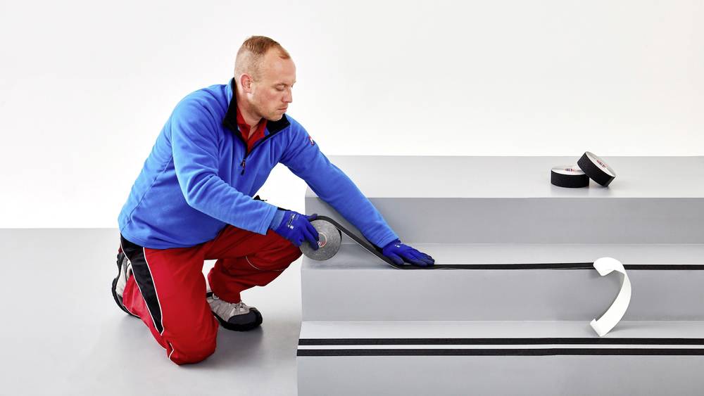 Mężczyzna klęczy i nakłada antypoślizgowe paski na stopień schodów. Ma na sobie ubranie robocze i rękawice ochronne.