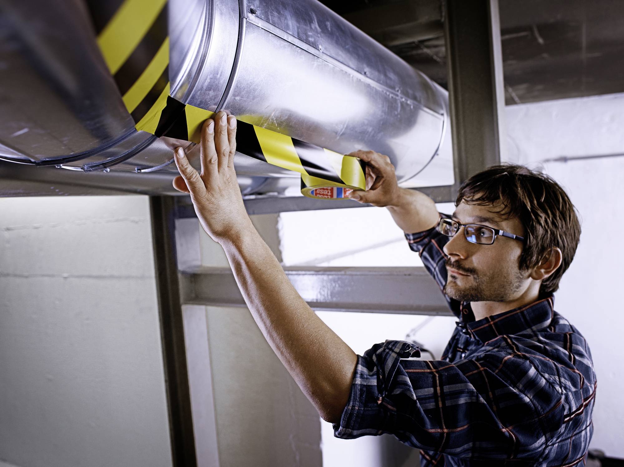 Mężczyzna sprawdza przewód wentylacyjny i nakłada na niego żółtą taśmę ostrzegawczą. Ma na sobie okulary i kraciastą koszulę.
