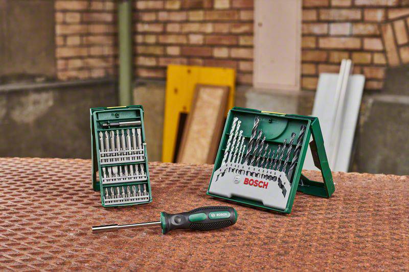 Zestawy wierteł i śrubokrętów firmy Bosch ułożone na stole, w tle ściana z cegły i materiały budowlane.