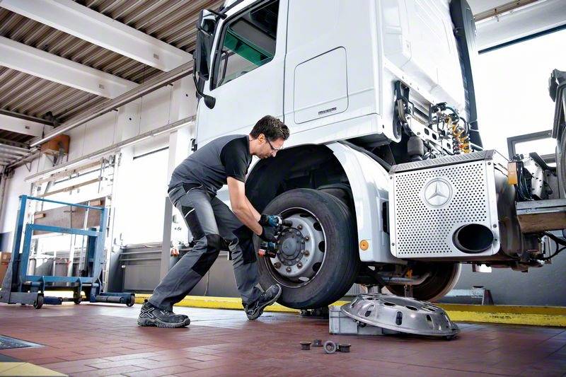 Mechanik wymienia oponę w ciężarówce w warsztacie. Ciężarówka jest podniesiona na podnośniku, a mechanik ma na rękach rękawice robocze.