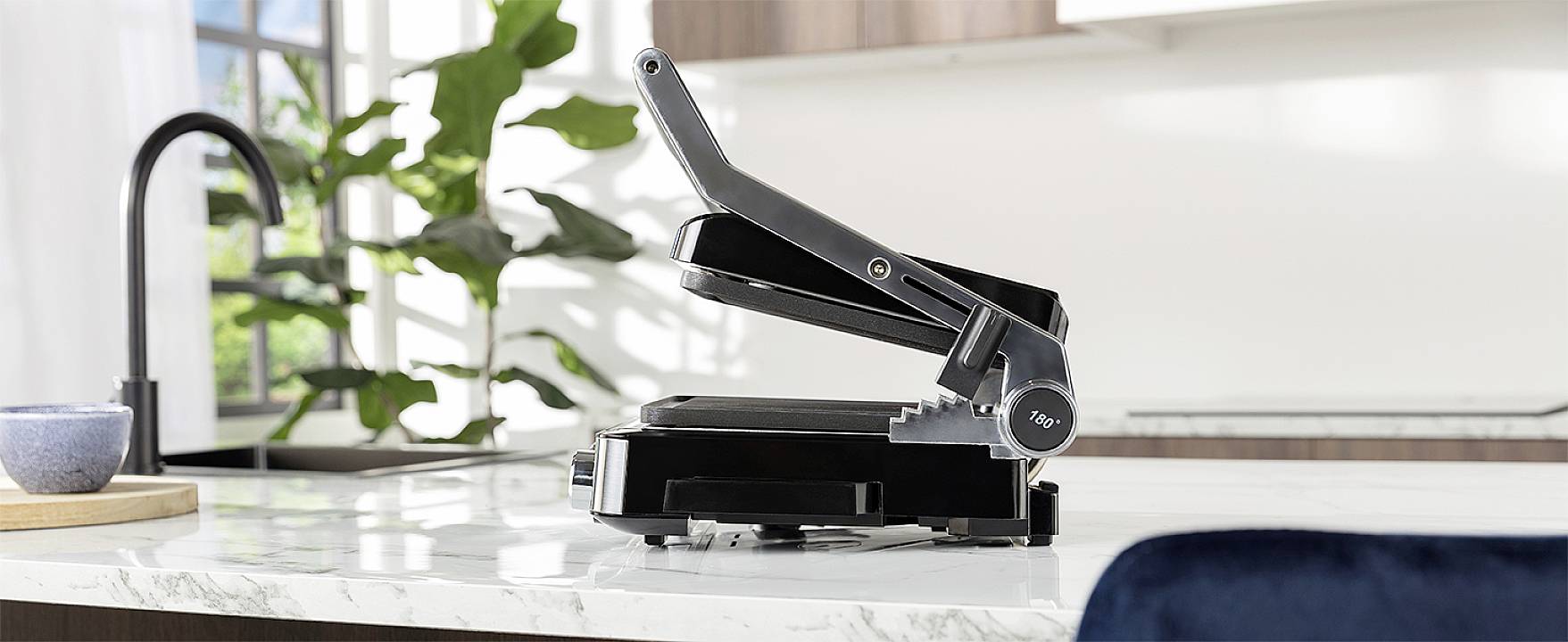 Czarny toster do kanapek stoi na białym blacie kuchennym przed oknem. Po lewej stronie obrazu widać roślinę.