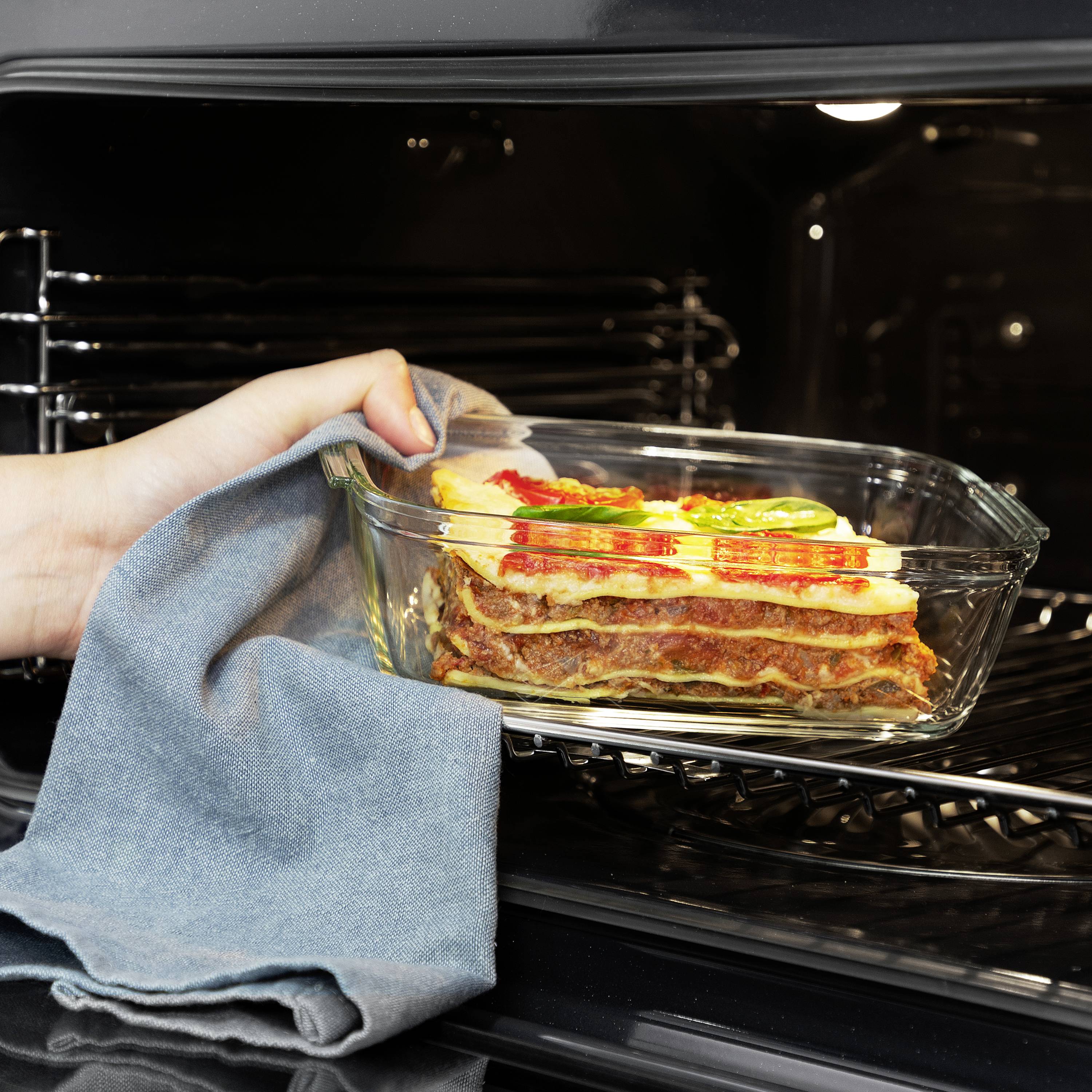 Ręka wyjmuje lasagne w przezroczystej żaroodpornej formie z piekarnika.