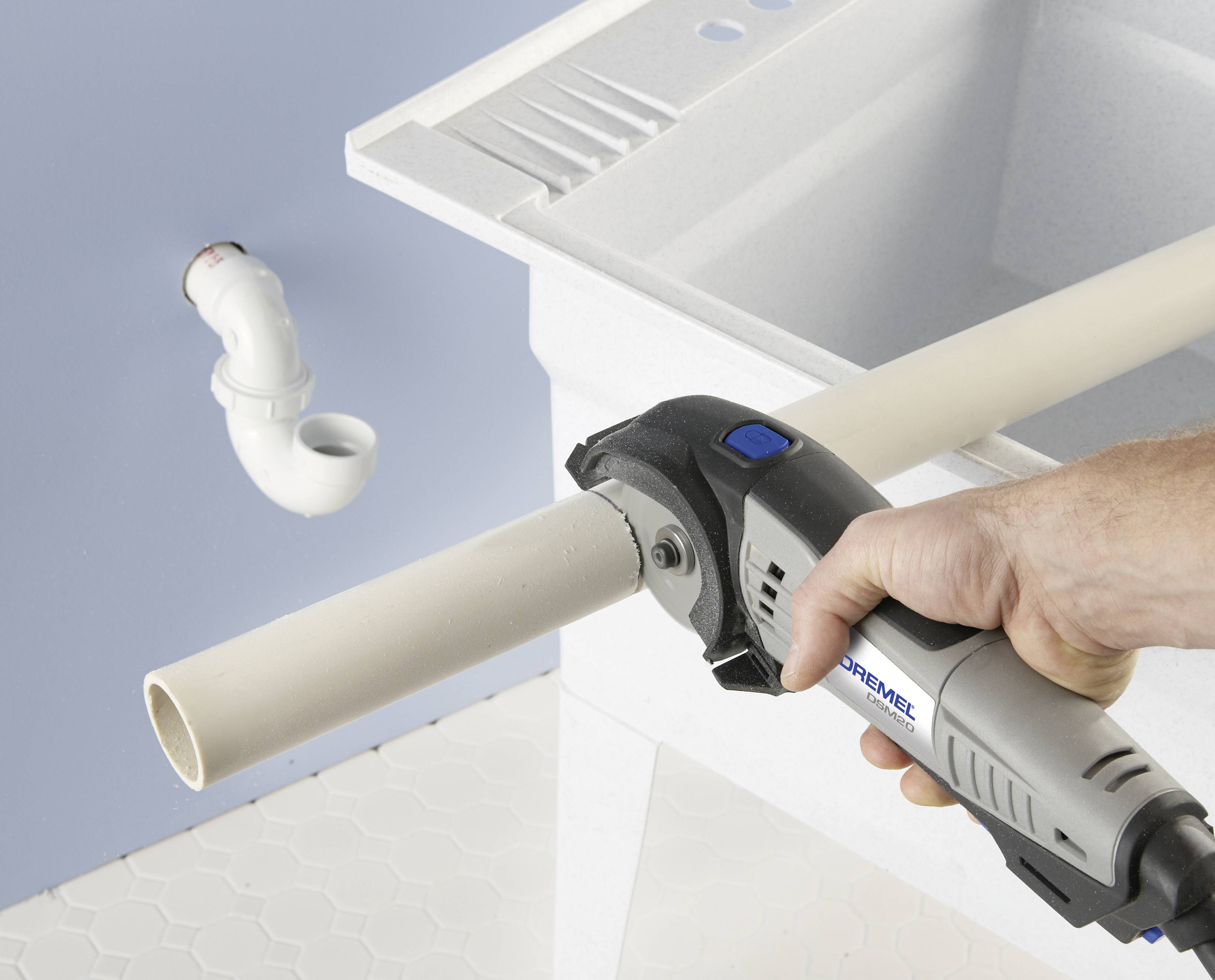 Ręka trzyma elektronarzędzie i przecina białą rurę z tworzywa sztucznego przed zlewem. W tle widoczna jest rura odpływowa.