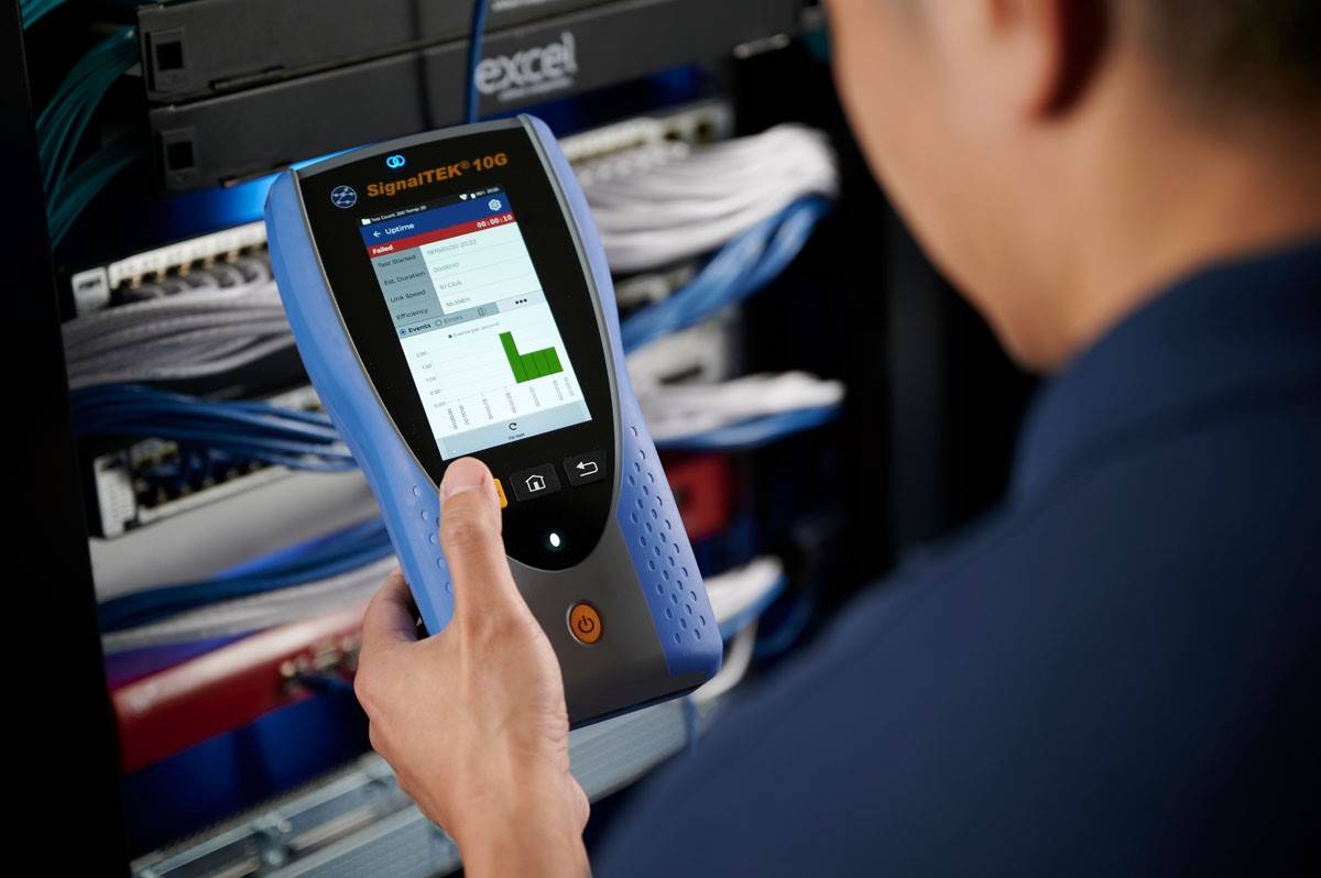 Osoba trzyma urządzenie SignaTEK 100 przed stosem serwerów sieciowych. Wyświetlacz pokazuje wykres słupkowy z danymi sieciowymi.
