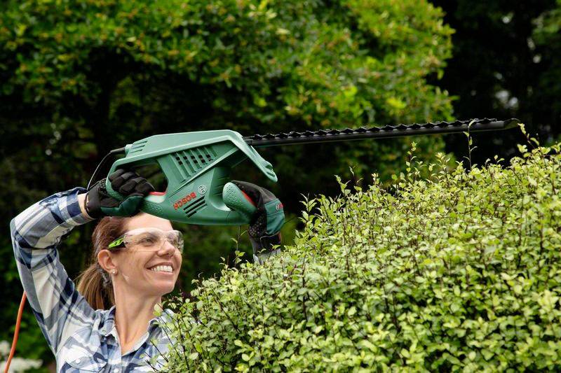 Kobieta promieniście uśmiechnięta przycina gęstą żywopłot elektrycznymi nożycami do żywopłotu. Ma na sobie okulary ochronne i rękawice w zielonym ogrodzie.