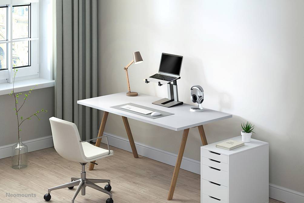 Nowoczesne biurko z podstawką na laptopa, lampą i słuchawkami. Biała szuflada obok, jasne otoczenie z oknem i roślinami.